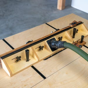 May include: A wooden woodworking jig with a black and green hose attached. The jig has a light wood finish with black accents and adjustable knobs. The hose is green with a black pattern. The jig is on a light wood surface.