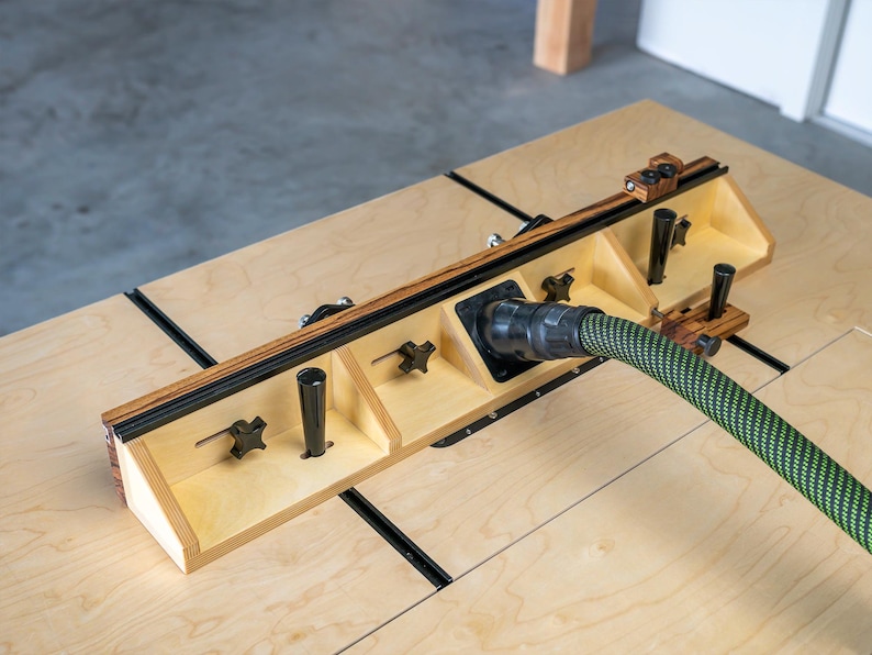 May include: A wooden woodworking jig with a black and green hose attached. The jig has a light wood finish with black accents and adjustable knobs. The hose is green with a black pattern. The jig is on a light wood surface.