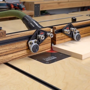 May include: A woodworking router table with a wooden fence and black hardware. A light-colored wooden board is being fed through the router. A green hose is connected to the router for dust collection.