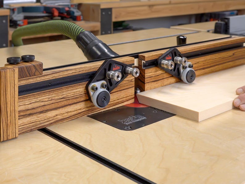 May include: A woodworking router table with a wooden fence and black hardware. A light-colored wooden board is being fed through the router. A green hose is connected to the router for dust collection.