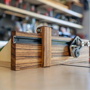 May include: A wooden woodworking jig with a dark wood grain finish. The jig features a sliding mechanism with metal tracks and adjustable components. The design includes a vertical support and a base, likely for precise cutting or shaping of wood.