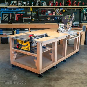 May include: A large wooden workbench on casters featuring a yellow DeWalt table saw integrated into the left side and a green and black Festool miter saw mounted on the right side. The workbench has a light wood finish and an open lower shelf.