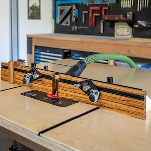 May include: A woodworking router table fence with a zebrawood pattern. The fence has black rails and adjustable components. A green hose is connected to the table, and the table is on a light-colored surface.