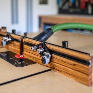 May include: A woodworking router table with a wooden fence and a green hose for dust collection. The table features a black base and silver hardware. The wood has a striped pattern.