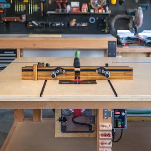 May include: A woodworking router table with a wooden fence and black metal components. The table is made of light-colored wood and sits in a workshop setting. The router table is designed for precision woodworking tasks.