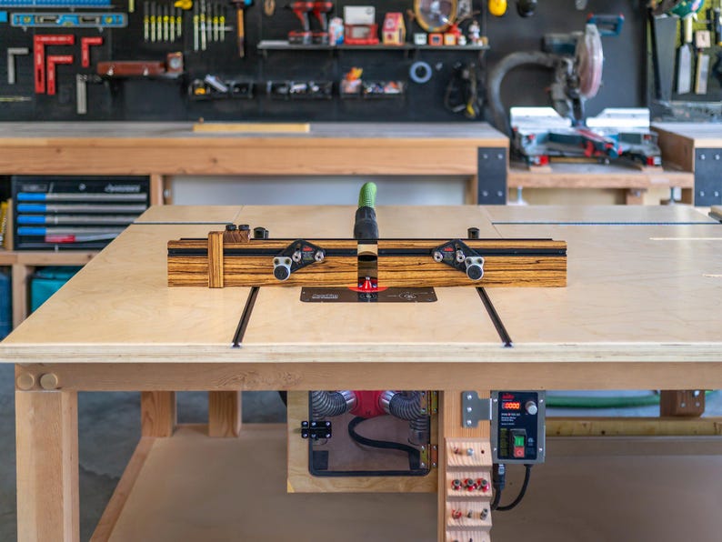 May include: A woodworking router table with a wooden fence and black metal components. The table is made of light-colored wood and sits in a workshop setting. The router table is designed for precision woodworking tasks.