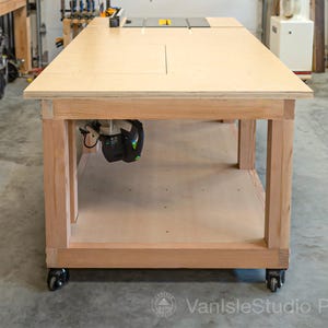 May include: A large, light-colored wooden workbench with a router mounted underneath. The workbench has a smooth, flat surface and is supported by sturdy wooden legs. It sits on wheels for mobility, and is designed for woodworking projects.