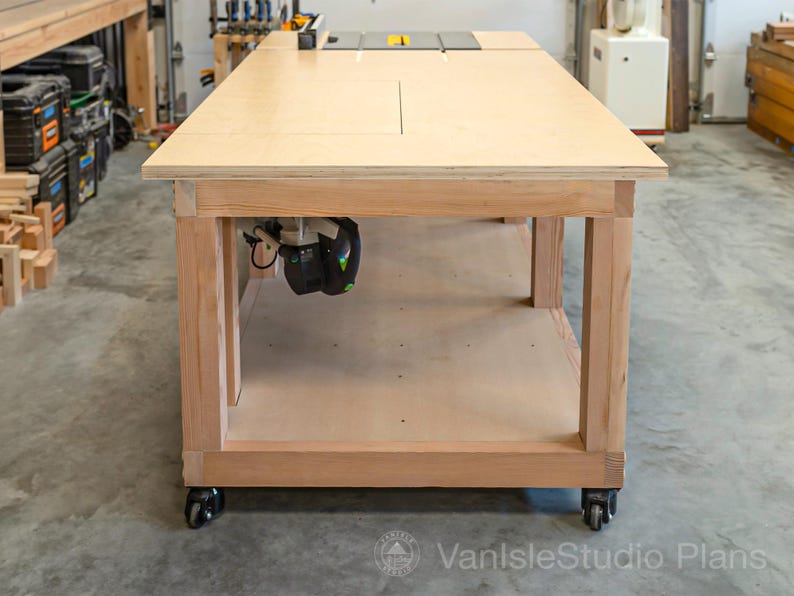 May include: A large, light-colored wooden workbench with a router mounted underneath. The workbench has a smooth, flat surface and is supported by sturdy wooden legs. It sits on wheels for mobility, and is designed for woodworking projects.