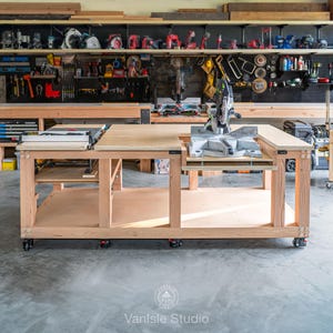 May include: A large, light-coloured wooden workbench on wheels featuring a built-in table saw on the left and a mitre saw integrated into the right side. The structure includes a lower storage shelf and a flat, expansive plywood work surface.