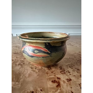 May include: A round ceramic bowl featuring a mottled glaze in shades of olive green, charcoal black, and burnt orange. The vessel has a wide, slightly flared rim and a smooth, curved body with a visible transition between the colors.