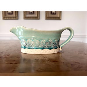 May include: A ceramic gravy boat with a turquoise glaze and a textured, wave-like design. The base is a creamy white, and the handle curves gracefully. The boat is on a wooden surface.