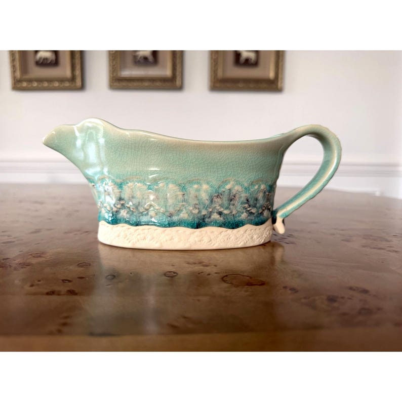 May include: A ceramic gravy boat with a turquoise glaze and a textured, wave-like design. The base is a creamy white, and the handle curves gracefully. The boat is on a wooden surface.