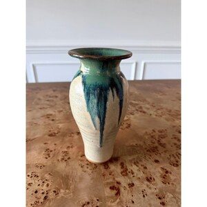 May include: A ceramic vase with a flared rim and a tapered body. The base is a speckled cream color, while the upper portion features a vibrant green and deep blue glaze that drips downward, creating a textured, layered appearance.