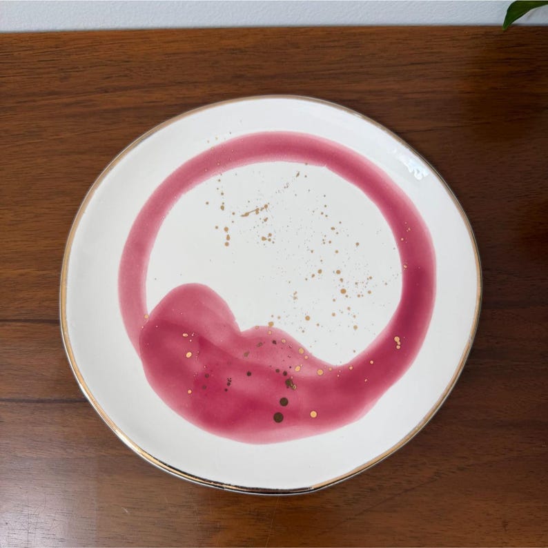 Puede incluir: Un plato de cer&aacute;mica blanco con borde dorado y un dise&ntilde;o abstracto audaz en tonos rojos y dorados. El dise&ntilde;o presenta una forma grande y curva y motas doradas dispersas, creando una est&eacute;tica moderna y art&iacute;stica.