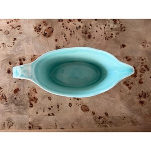 May include: A light blue ceramic serving dish with a long, oval shape and two handles. The interior of the dish is a solid light blue, while the exterior has a slightly textured finish. The dish is set against a wooden surface.