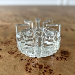 May include: A clear glass candle holder with a circular design. The candle holder has multiple cylindrical sections and a textured surface. The design is reminiscent of a crown or a starburst. The glass is transparent, allowing light to pass through.