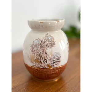 May include: A white ceramic vase with a brown and gold floral design. The vase has a rounded body and a slightly flared rim.