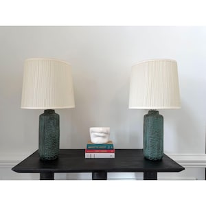 May include: Two matching green ceramic lamps with cream-colored pleated shades sit on a black table. A small white sculpture and a stack of books are between the lamps. The lamps have a textured, patterned design.