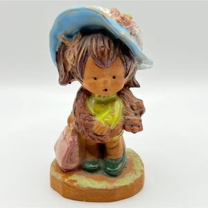 May include: A ceramic figurine of a child wearing a blue hat with pink flowers and a green top. The figurine is holding a pink purse and is standing on a brown base. The child has brown hair and green boots.