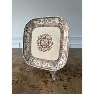 May include: A square ceramic plate with rounded corners, featuring a cream-colored base and intricate brown transferware patterns. The center displays a circular floral motif, while the border is decorated with detailed botanical and bird imagery.