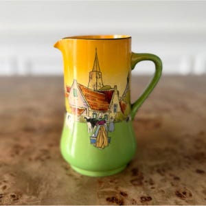 May include: A vintage ceramic pitcher with a vibrant design. The top half is a gradient of yellow to orange, depicting a village scene with a church. The bottom half is a bright green, and the handle is green. A charming decorative piece.