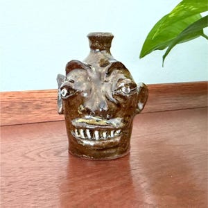 May include: A brown glazed ceramic face jug with a narrow neck and a wide opening. The jug features a sculpted human-like face with prominent ears, wide eyes, and a mouth showing a row of white teeth. The surface has a glossy, mottled finish.
