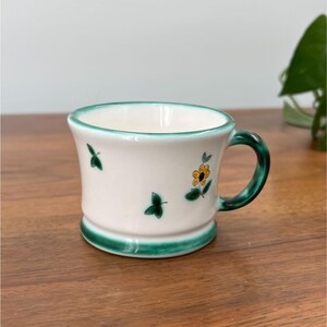 May include: White ceramic mug with a green handle, rim, and base. The mug is decorated with small green leaf and yellow flower designs. The mug is sitting on a wooden surface.