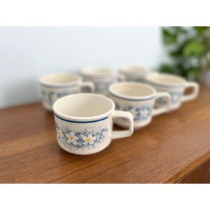 May include: A set of six ceramic mugs with a cream-colored base. Each mug features a blue band near the rim and a floral pattern of white and blue flowers. The mugs have a simple handle design and are arranged on a wooden surface.