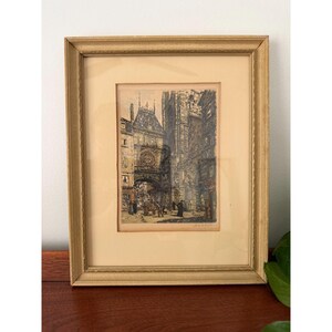 May include: A framed etching depicting a historic European street scene with a large stone archway, tall buildings, and small figures walking below. The artwork is presented in a light-colored wooden frame with a wide, cream-colored matting border.