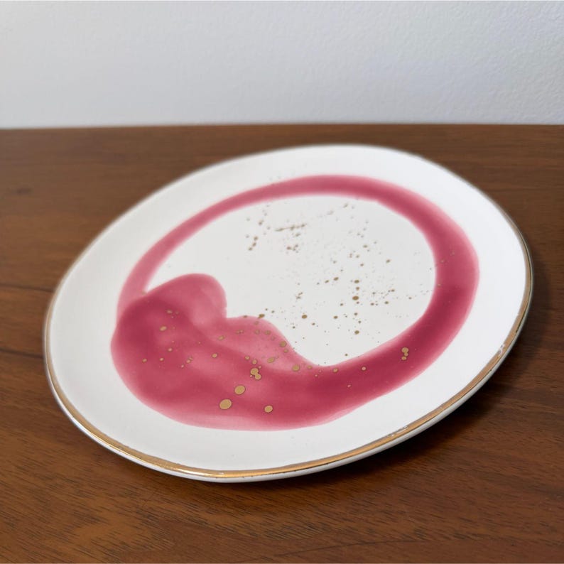 Puede incluir: Plato de cer&aacute;mica blanca con borde dorado y dise&ntilde;o abstracto burdeos. El dise&ntilde;o incluye una forma circular y una salpicadura de pintura burdeos con motas doradas. El plato est&aacute; sobre una superficie de madera.