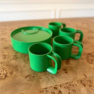 May include: A set of four bright green plastic mugs with square-shaped handles and a matching circular green container with a lid. The mugs are arranged in a cluster next to the container, all featuring a smooth, solid-colored finish.