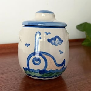 May include: A cream-colored ceramic jar with a matching lid, featuring a hand-painted blue whale design swimming in green and blue waves. The scene includes small blue birds and a cloud above the whale, with blue trim on the lid and rim.