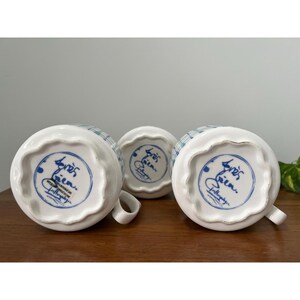 May include: Three white ceramic mugs with a blue and green plaid pattern. The bottom of each mug features a blue circular design with script and the words "Anthropologie". Each mug has a small handle.