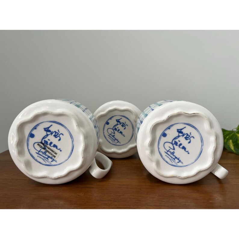 May include: Three white ceramic mugs with a blue and green plaid pattern. The bottom of each mug features a blue circular design with script and the words "Anthropologie". Each mug has a small handle.