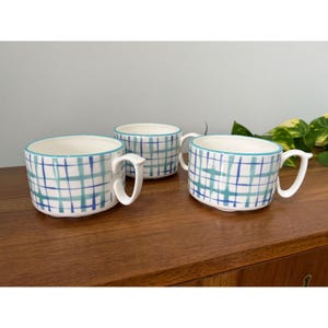 May include: Three white ceramic mugs with a blue and teal plaid pattern. Each mug has a white handle and a teal rim. The mugs are arranged on a wooden surface.