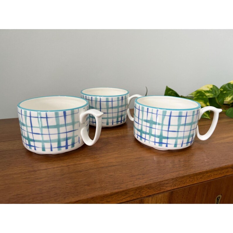 May include: Three white ceramic mugs with a blue and teal plaid pattern. Each mug has a white handle and a teal rim. The mugs are arranged on a wooden surface.