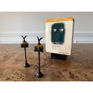 May include: A set of two miniature black lamp posts with orange globes topped by bat figures, accompanied by a Department 56 Village Accessories box labeled Bat Lights (Set of 2) with Halloween branding and an image of the lamps.