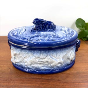 May include: A blue and white ceramic lidded dish with a rabbit figurine on top. The dish has a round shape with a detailed relief pattern of foliage and a two-tone color scheme. The lid and base are a deep blue, while the sides feature a white and blue gradient.