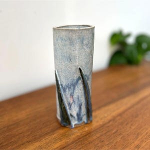 May include: A tall, cylindrical ceramic vase with a textured, mottled gray and blue glaze. The vase features two dark blue, curved, decorative elements extending from the base. The vase is displayed on a wooden surface.