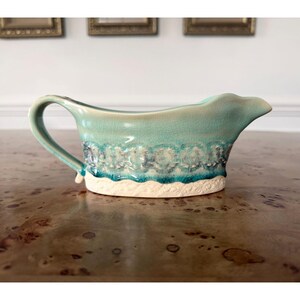 May include: A turquoise ceramic gravy boat with a handle and spout. The boat features a textured design with white and blue accents along the rim and base. The gravy boat is on a wooden surface.