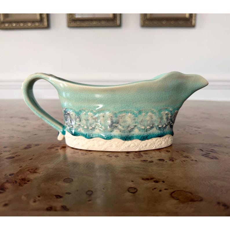 May include: A turquoise ceramic gravy boat with a handle and spout. The boat features a textured design with white and blue accents along the rim and base. The gravy boat is on a wooden surface.