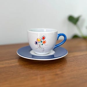 May include: A white ceramic teacup and saucer set. The cup has a blue handle and a blue rimmed saucer. The cup features a colorful floral design with a pair of hands holding the flowers. The set is on a wooden surface.