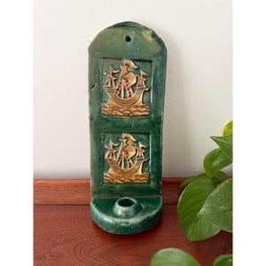 May include: A vertical, dark green glazed ceramic wall sconce featuring two stacked, embossed square panels. Each panel depicts a stylized sailing ship in a warm, light brown tone. The base includes a circular candle holder cup.