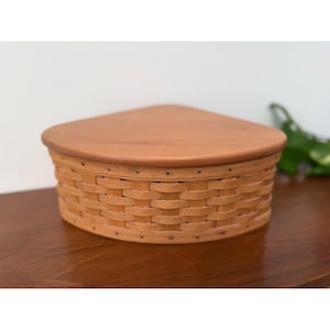 May include: A light brown, triangular-shaped basket with a matching wooden lid. The basket is made of woven strips of wood, and the lid is rounded. The basket is sitting on a wooden surface.