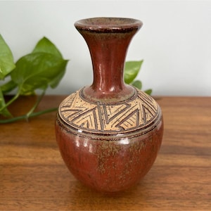 May include: A handcrafted, reddish-brown ceramic vase with a flared rim and a rounded body. The vase features a geometric pattern in tan and brown around the neck. The vase is displayed on a wooden surface, showcasing its earthy tones and artisanal design.