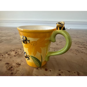 May include: A yellow ceramic mug featuring painted sunflower motifs and three-dimensional bee figures. The handle is light green and shaped like a vine, topped with a small bee. The mug has a white interior and a tapered cylindrical body.