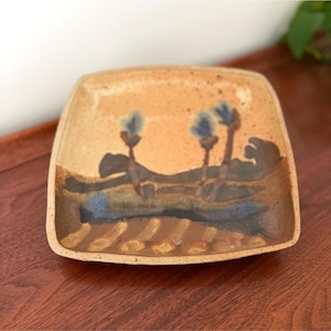 May include: A square ceramic plate with rounded corners, featuring a speckled beige surface. The center displays a hand-painted landscape with dark brown rolling hills, three blue-tipped vertical plant motifs, and a textured brown foreground.