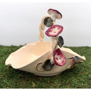 May include: Cream-colored ceramic basket with a twisted handle and floral accents. The basket features a scalloped rim and is adorned with stylized flowers in shades of red, white, and gray. A small hummingbird is perched on the side. A decorative piece for the home.