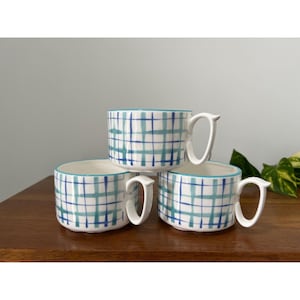 May include: Three white ceramic mugs with a blue and teal plaid pattern. Each mug has a curved handle and a teal rim. The mugs are stacked on a wooden surface.