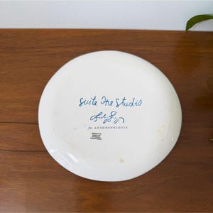 Puede incluir: Plato de cer&aacute;mica blanco con la inscripci&oacute;n azul "Suite One Studio" y una firma, "for ANTHROPOLOGIE". El plato es redondo con un borde ligeramente elevado. Sobre una superficie de madera marr&oacute;n.